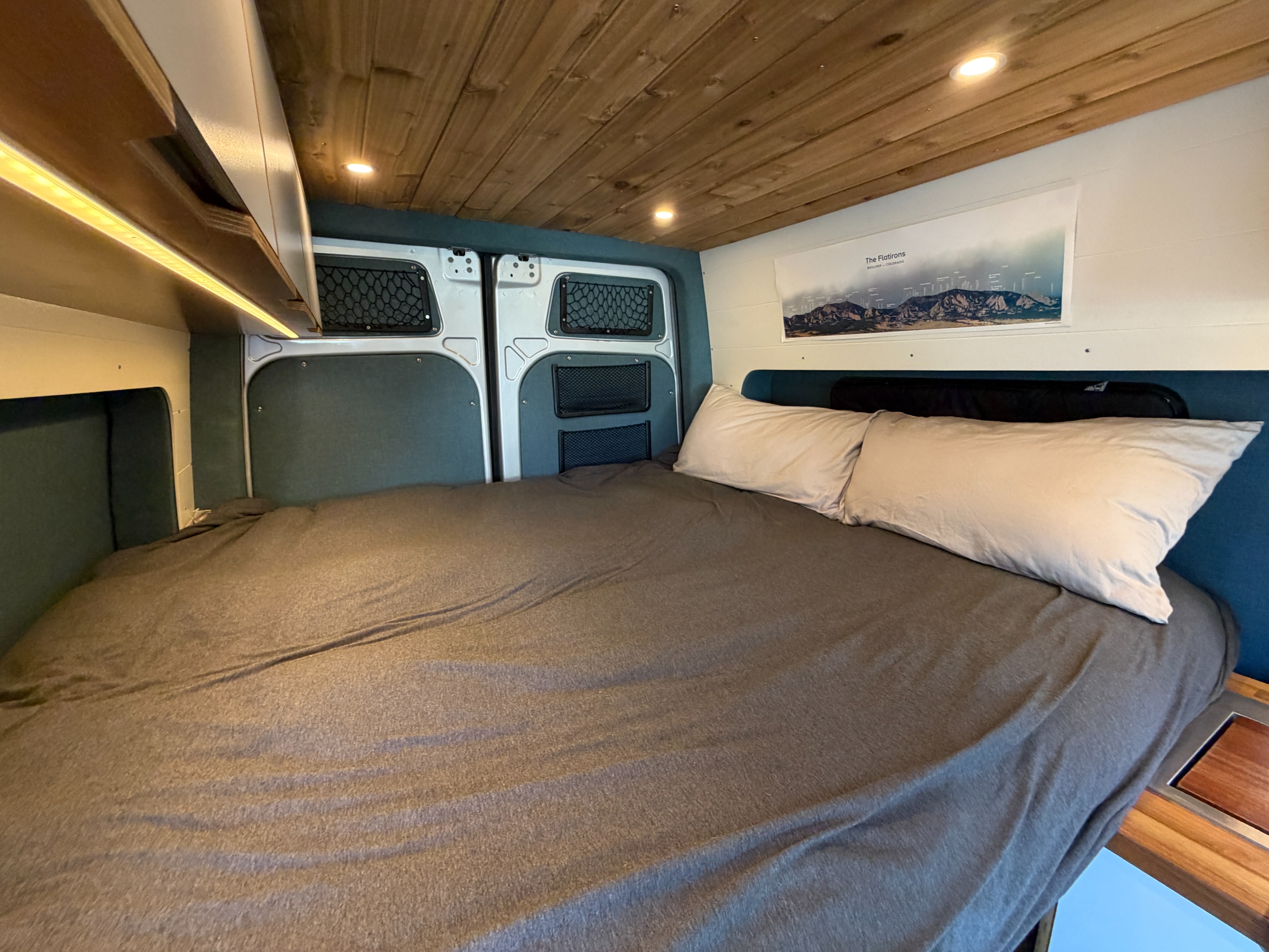 Inside an rv rental: a cozy van bedroom with a grey bed, pillows, wooden ceiling, lights, and back door storage.. Mercedes-Benz Sprinter 2019