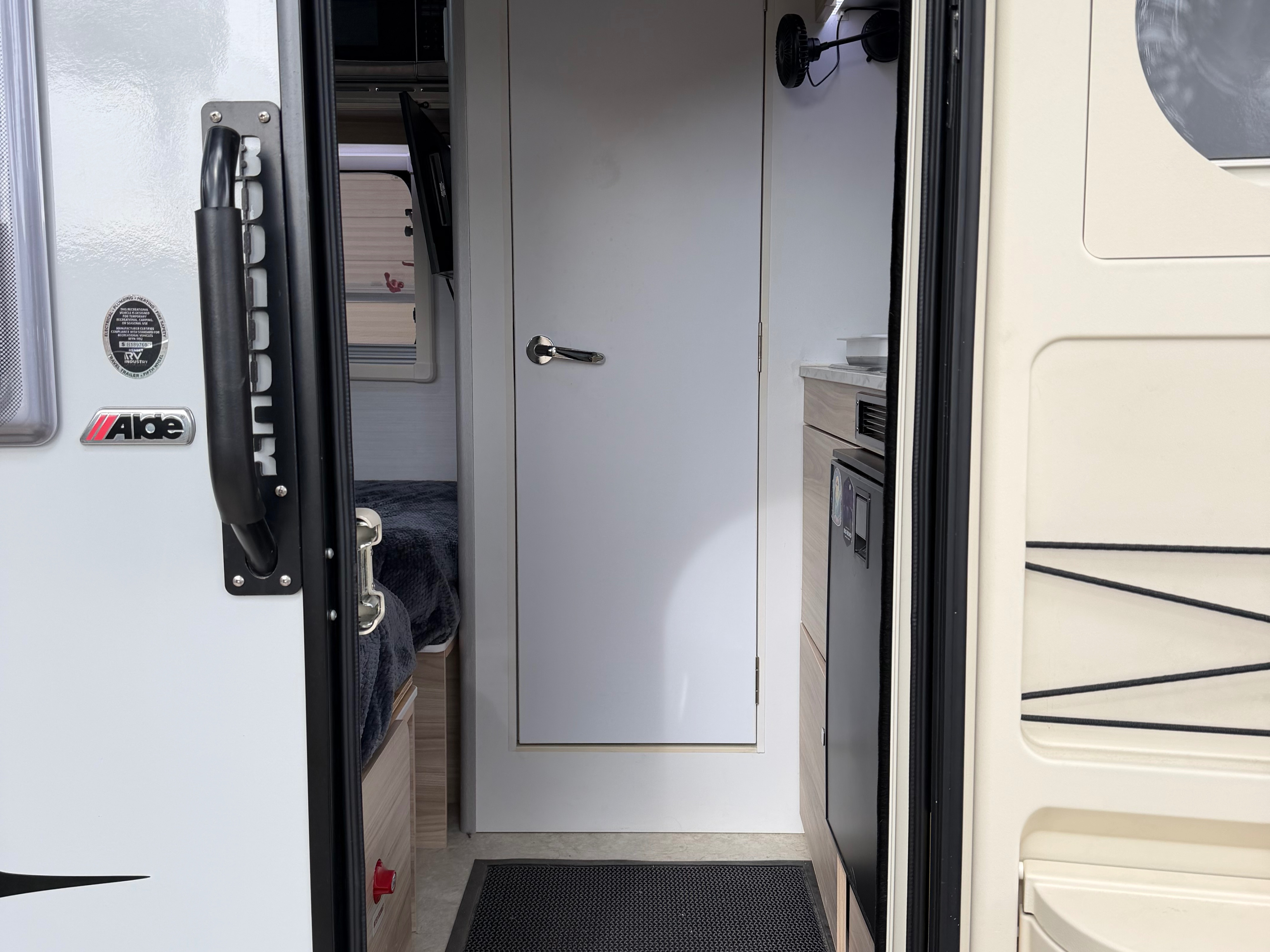 This RV rental interior features a cozy bedroom on the left, a white door, and a compact kitchen on the right with a fridge.. nuCamp T@B 2024