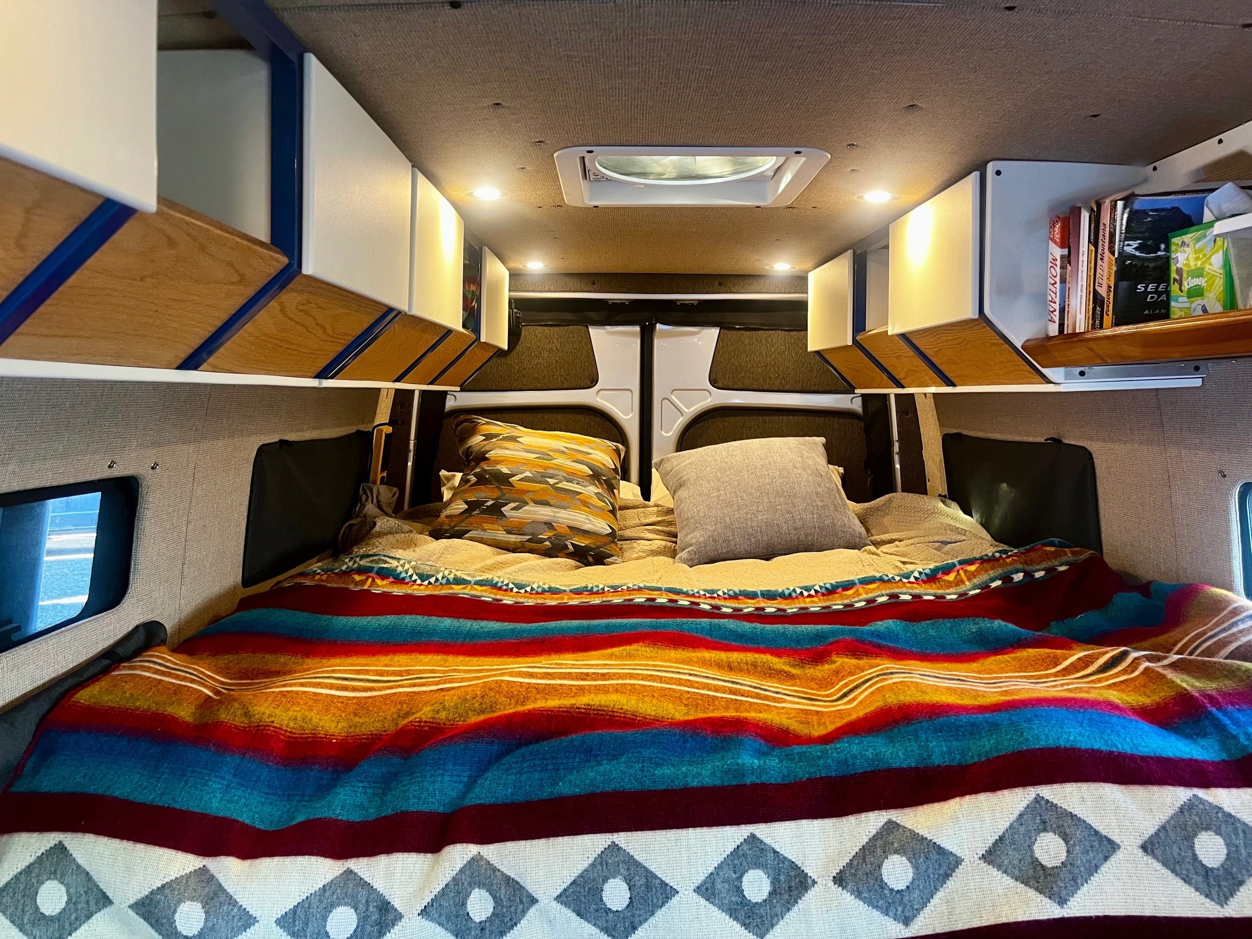This rv rental boasts a cozy sleeping area, featuring a vibrant blanket, pillows, and custom overhead cabinets for travel comfort.. Mercedes-Benz Sprinter 2019