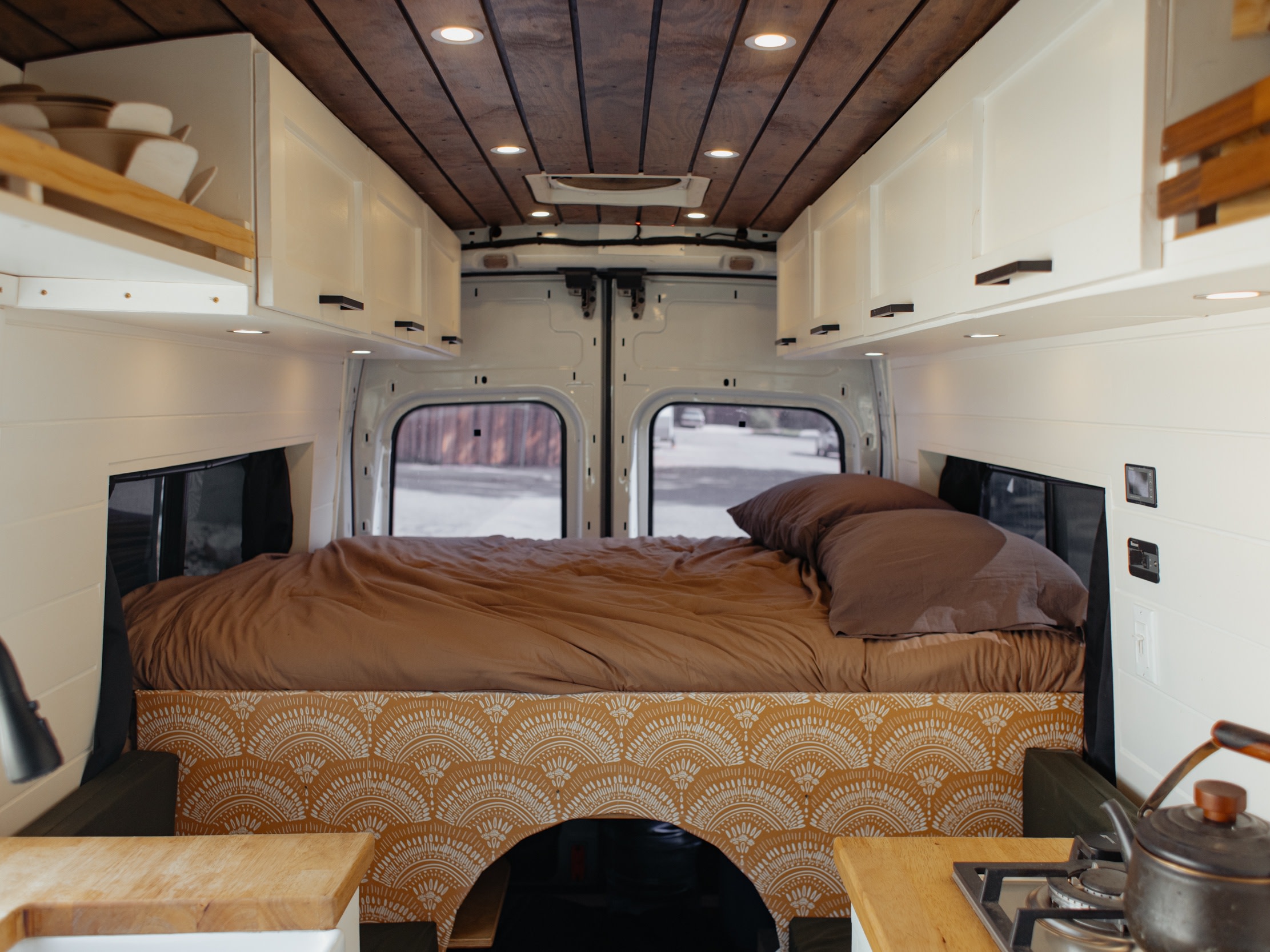 Cozy RV rental van interior with a bed in the rear, wood-paneled ceiling, white cabinets, and a compact kitchen counter with a stove.. Ford Transit 2015