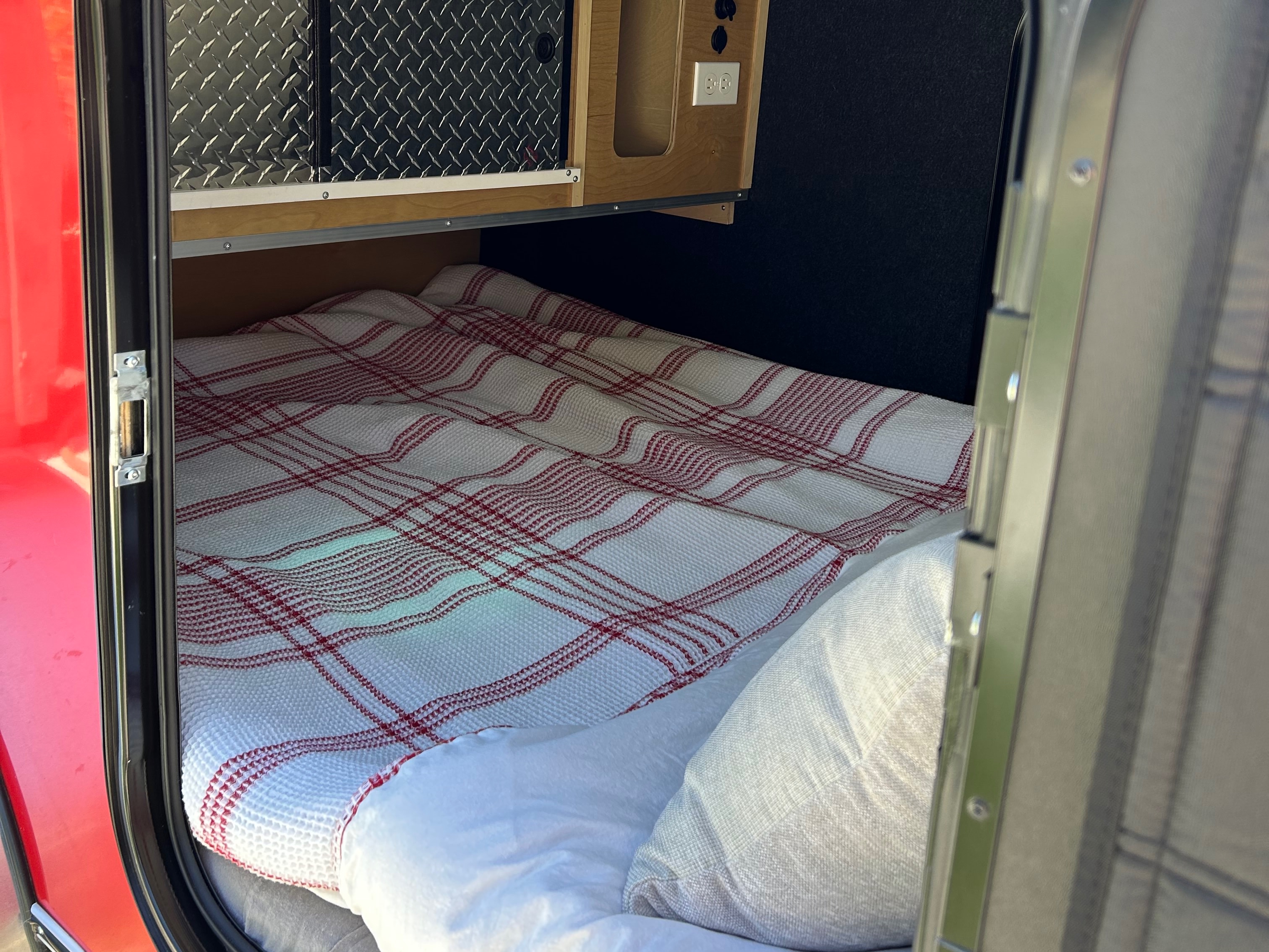 Inside a compact RV rental: a cozy bed with red plaid blanket, storage, and outlets for a comfortable trip.. American Teardrop NA 2019