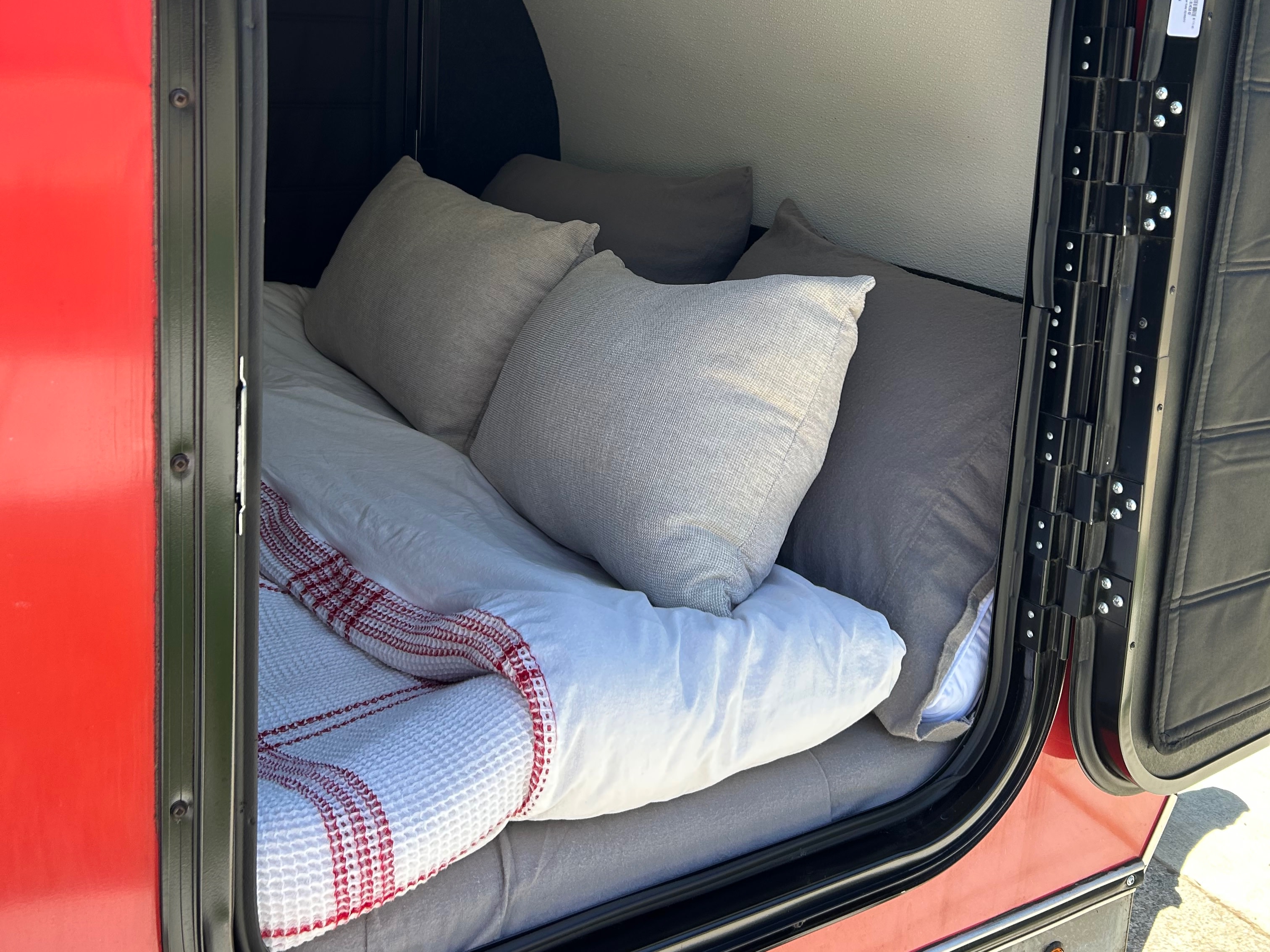 Inside this RV rental: a cozy camper bed setup with grey pillows, white duvet, and a striped blanket, ready for rest.. American Teardrop NA 2019