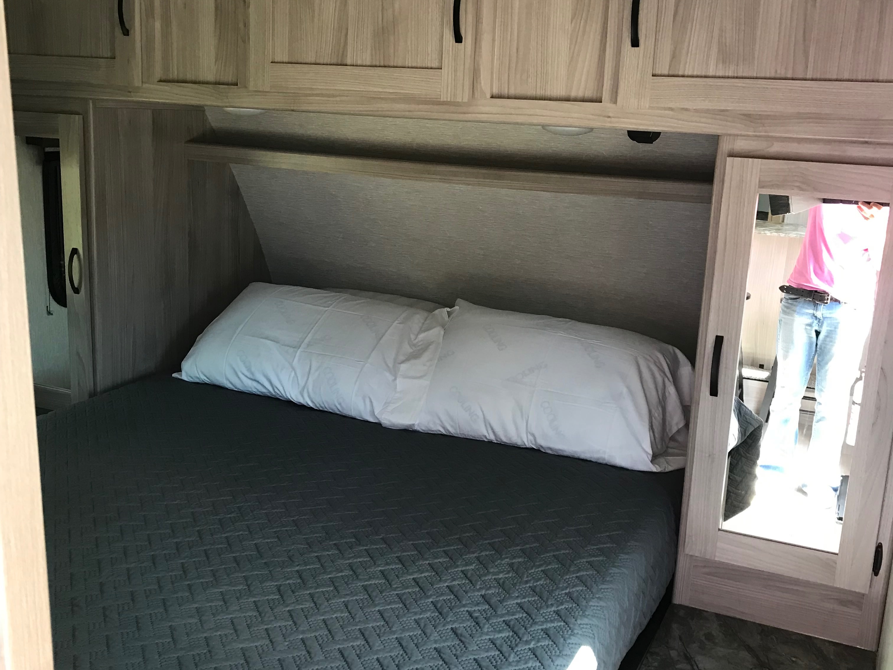 Inside an rv rental: a cozy bedroom with a dark gray quilted bedspread, pillows, and overhead storage. Person reflected in mirror.. Forest River Coachmen Northern Spirit Ultra Lite 2022