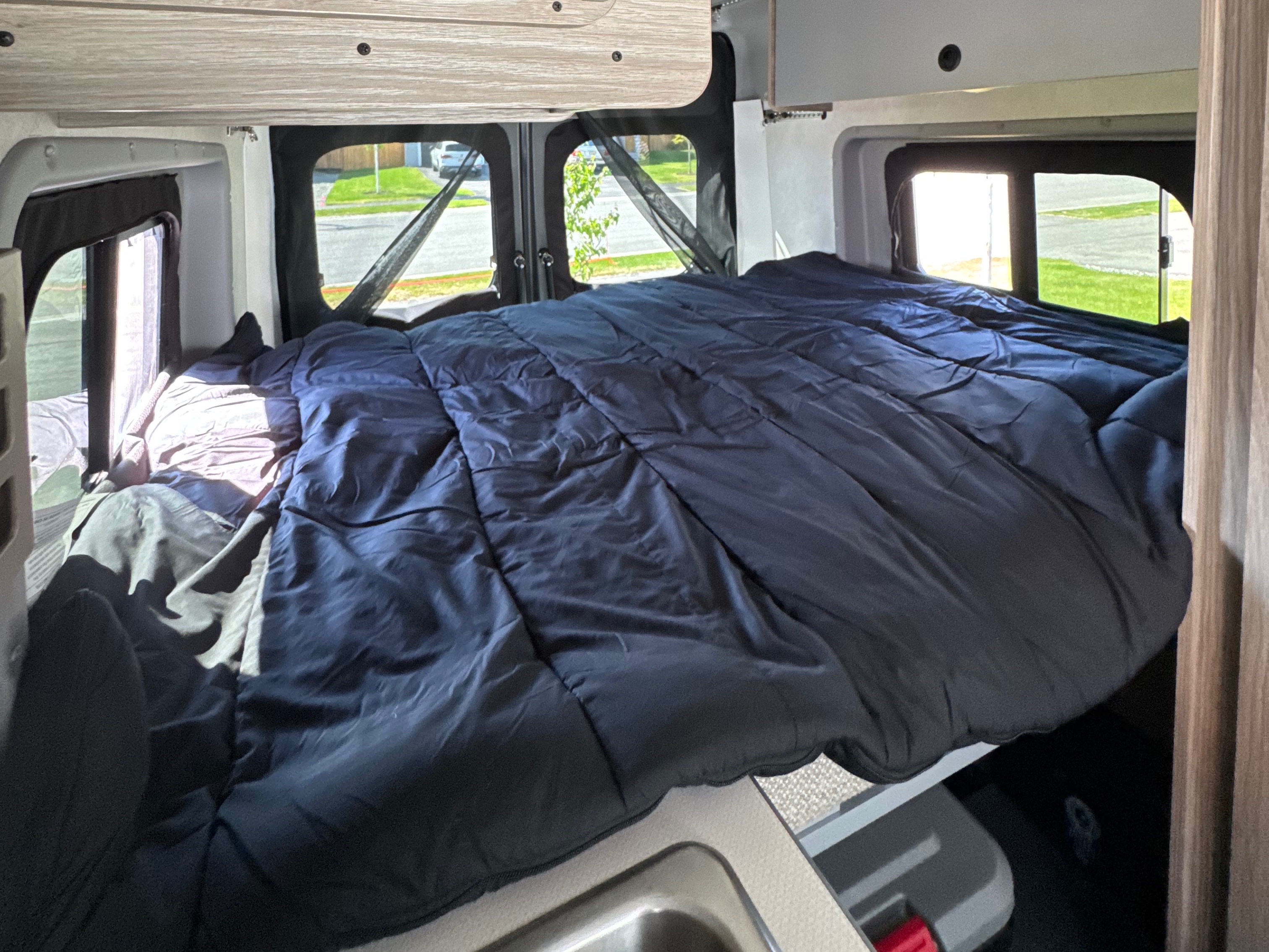 Cozy RV rental bed with a dark blue comforter, screens on windows, and a view of the street outside.. Winnebago Solis 59PX 2025
