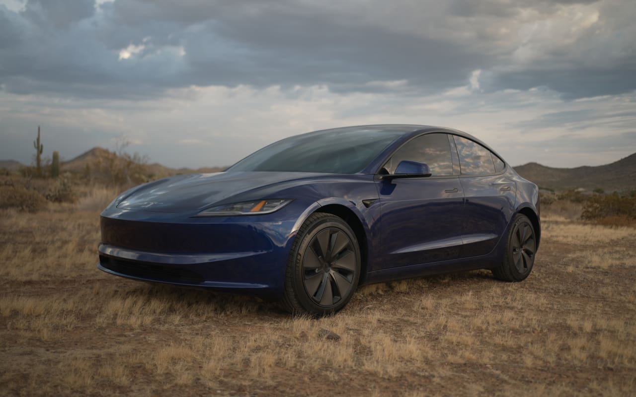 A dark blue Tesla electric car in a dry desert with mountains & saguaro cacti under a cloudy sky
