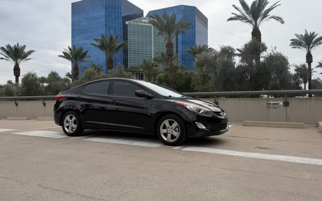 A black sedan is parked in a lot with palm trees and modern buildings