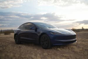 A blue Tesla Model 3 sits in a dry desert landscape under a cloudy sunset sky