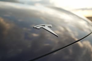 A close-up of a Tesla logo on a sleek car hood, reflecting clouds and golden light