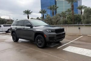 A dark grey Jeep Grand Cherokee L SUV parked on a wet lot, with palm trees and a blue building