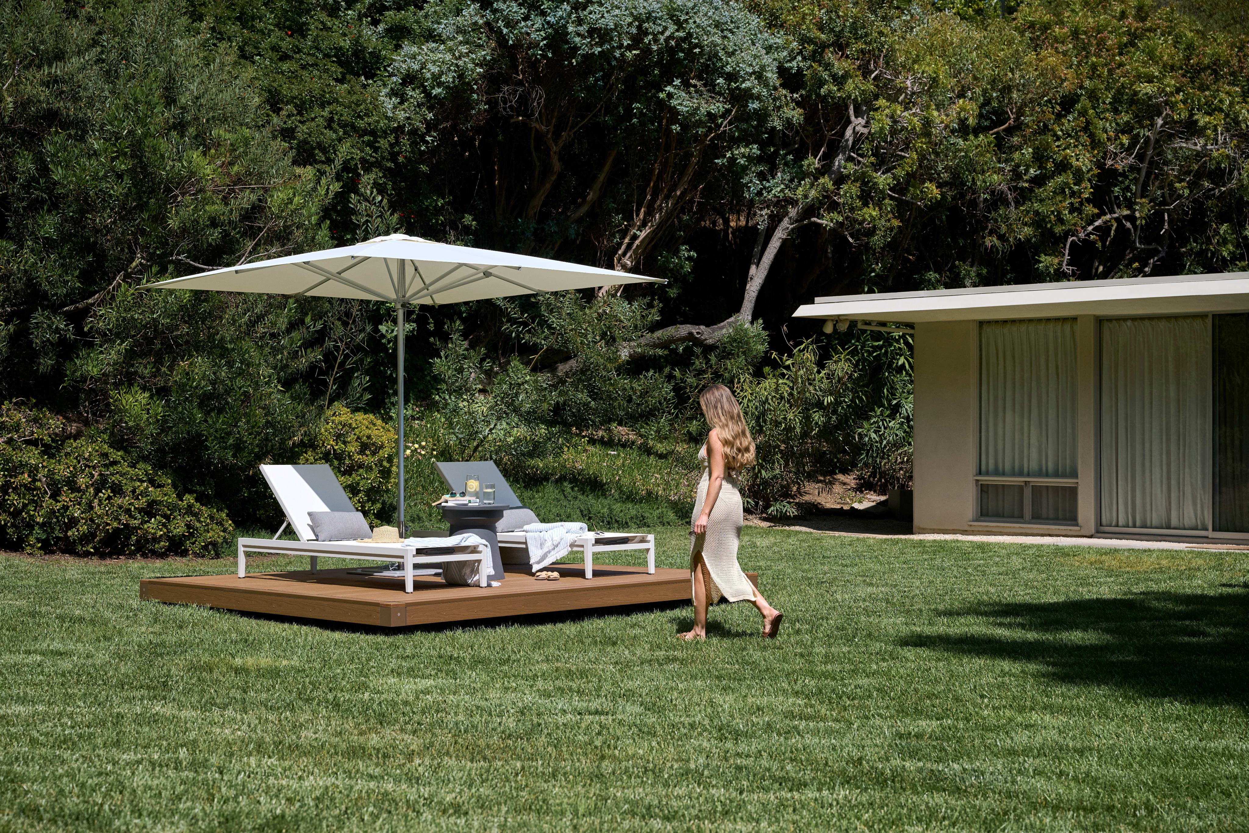 A woman walking toward an OS2 freestanding deck with loungers and a parasol on a sunlit lawn