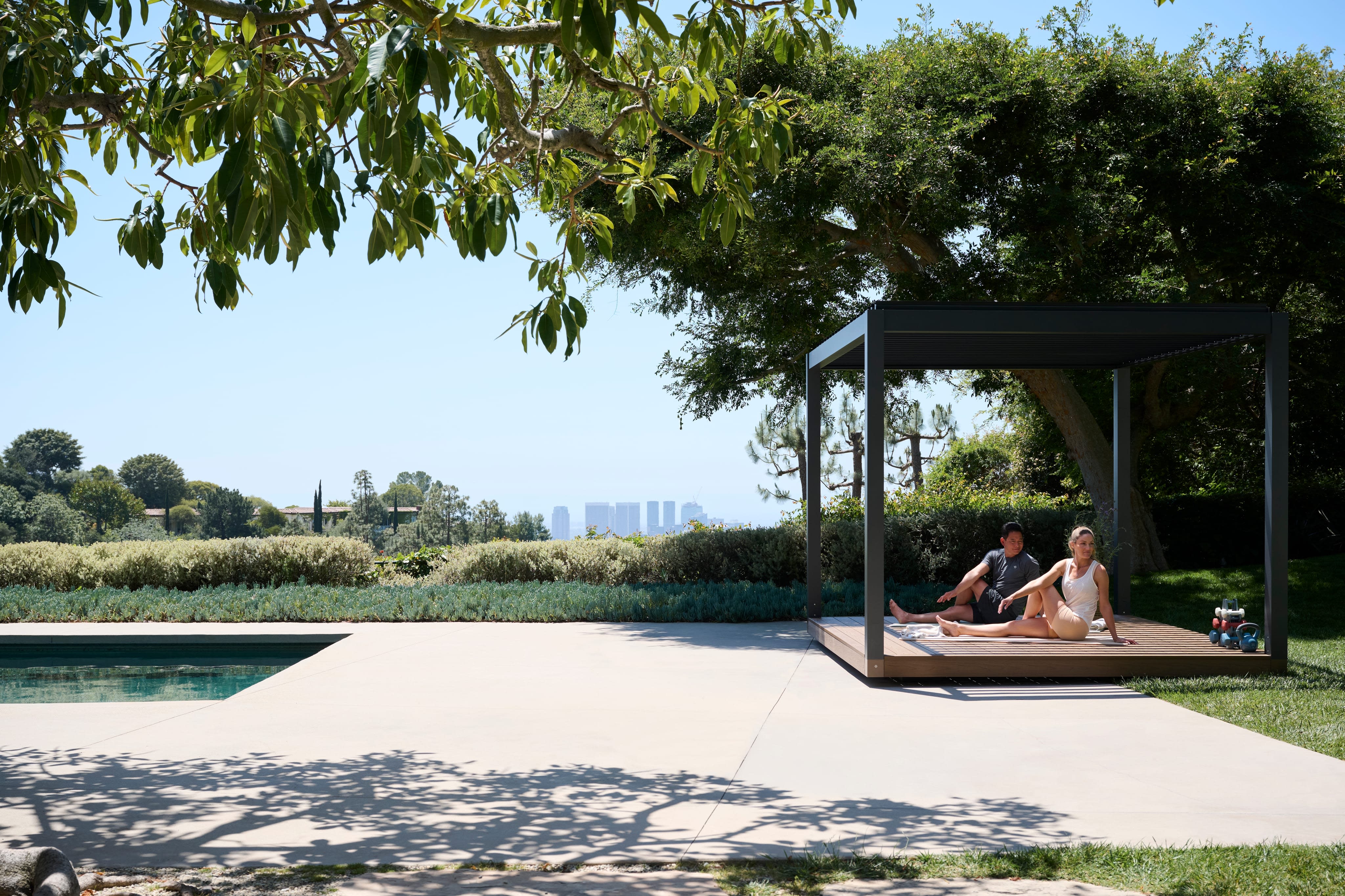 Two people stretching on an Outer Spaces pavilion deck beside a pool, with a city skyline beyond the hedge