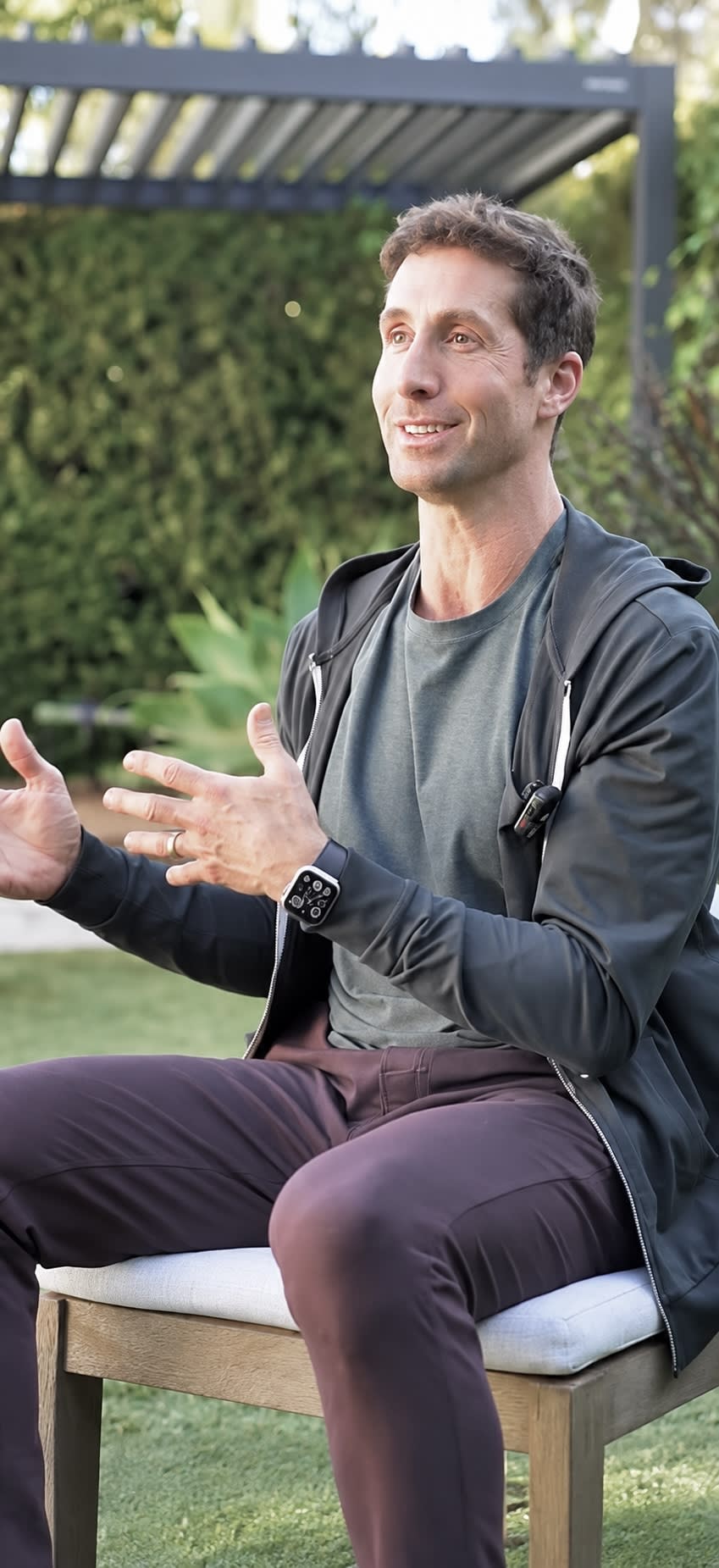 David talking and gesturing while seated in front of a louvered pergola