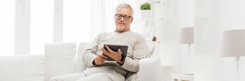 Image shows a man around 50 years old on his electronic tablet. He is wearing casual, light coloured clothing, wears glasses and is sitting on a sofa in a living room. He is looking at the tablet like he is reading on it.