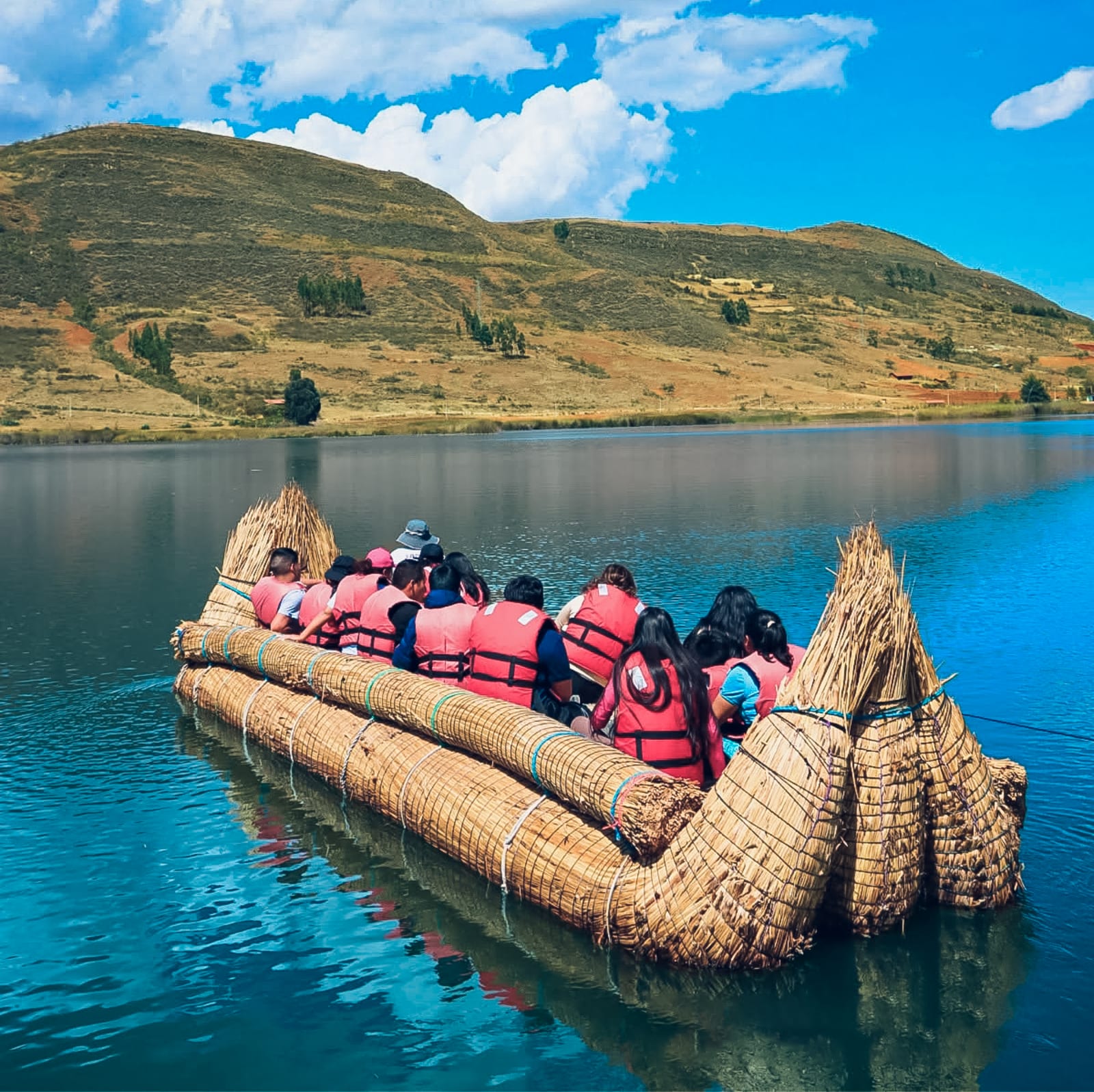 Un grupo de visitantes con chaleco salvavidas naranja cruza una laguna andina en una balsa de totora, con cerros secos al fondo