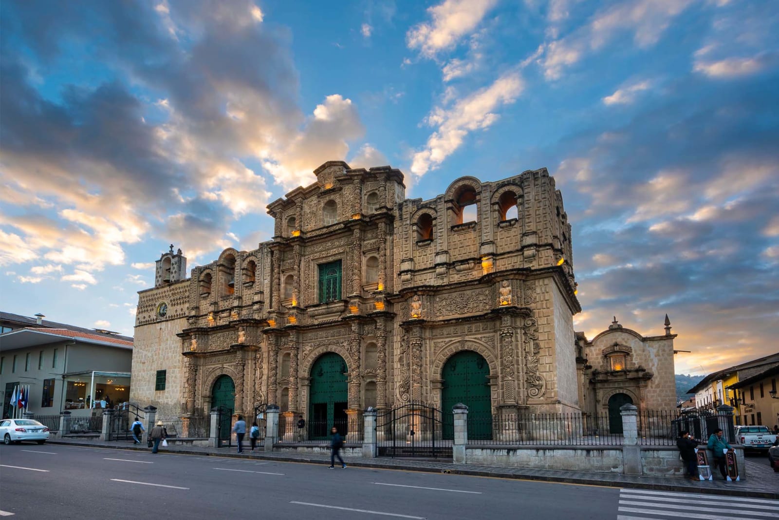 La catedral de Cajamarca al atardecer, con la fachada de piedra tallada iluminada y gente cruzando la Plaza de Armas