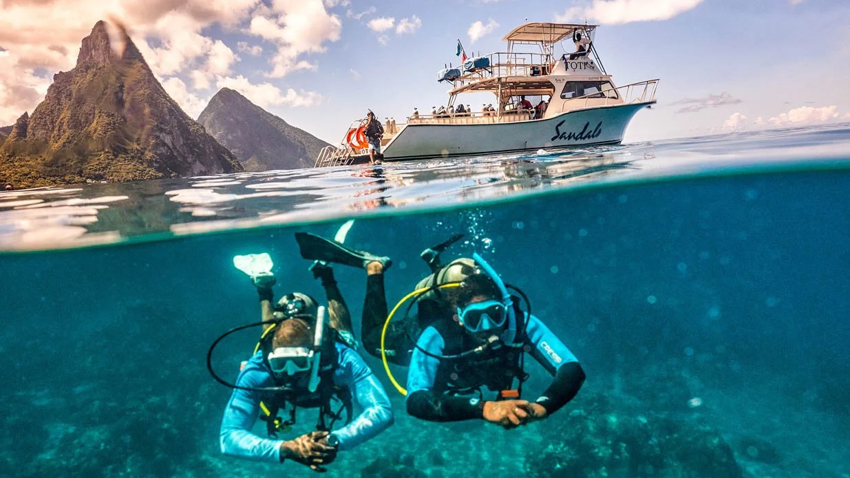 Two scuba divers swim underwater near a dive boat named "Sandals" in clear tropical waters