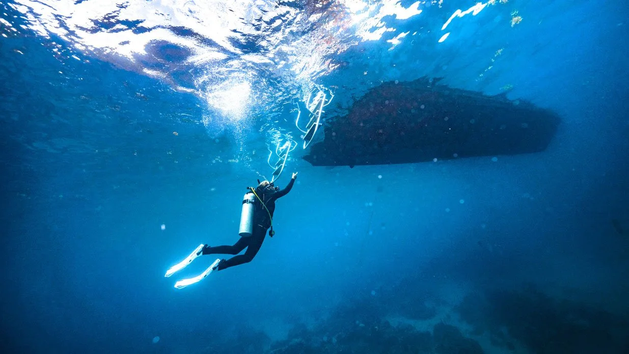 A scuba diver ascends toward the surface of the ocean, reaching for a boat overhead. 