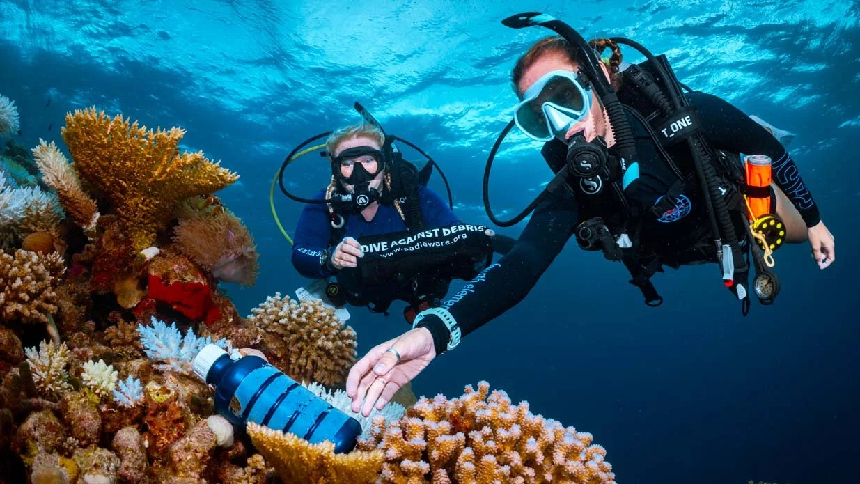 A scuba diver swims upward through clear blue water, reaching toward the hull of a boat above. 