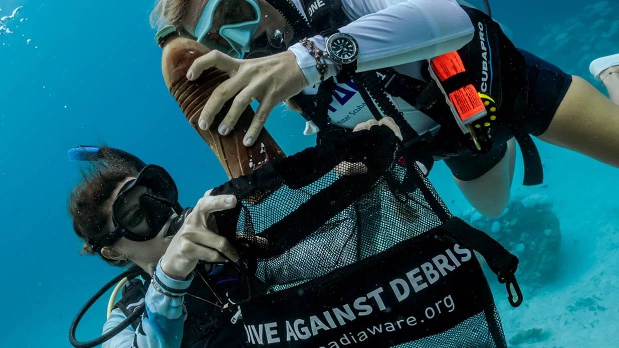 Two scuba divers underwater collecting marine debris in a black mesh bag labeled 'DIVE AGAINST DEBRIS'