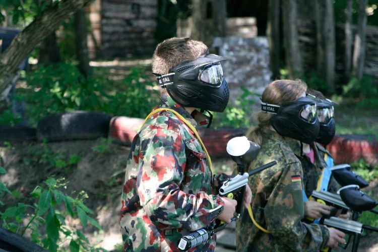 Trójka młodych graczy w paintball w cieniu drzew z okopami w tle