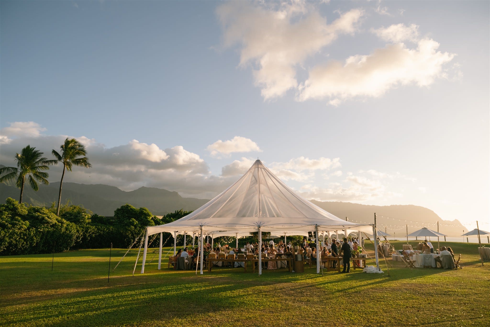 Tented wedding reception on a coastal lawn at golden hour, with palm trees and mountains behind