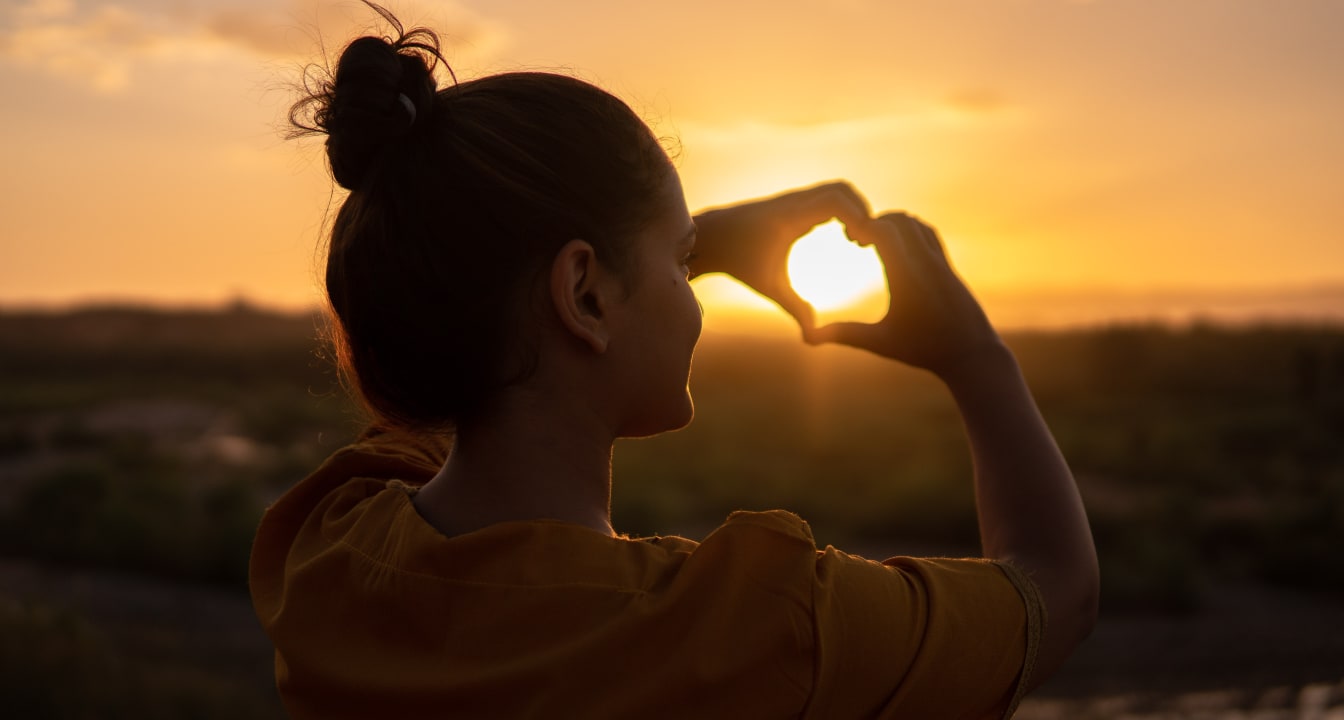 Woman making love heart shape with her hands in front of a setting sun