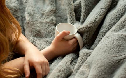 Student snuggles under a grey fur blanket with a mug in her hand.