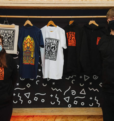 A man and women stand at either end of the photo. Both are wearing hoodies with the Keep Leeds Warm logo on. Keep Leeds Warm t-shirts are displayed behind them