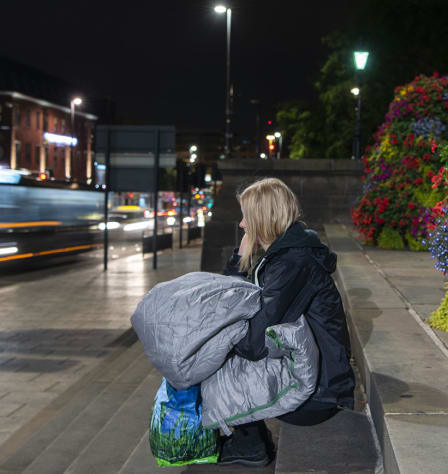 A woman sits on some steps. She has a sleeping bag in her lap. Cars speed off past her. Flowers can be seen behind her.