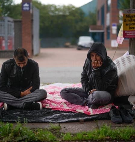 Two men sit on the floor with a blanket and plastic sheeting underneath them. One man is looking down, the other has a hood up and is resting his face on his chin.