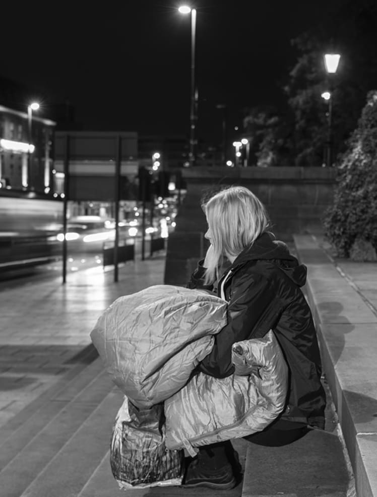 A woman sits on a set of steps. She is holding a sleeping bag and looking at the traffic passing by.