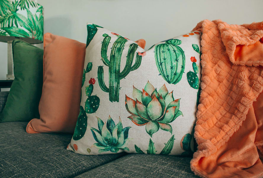 Orange and green sofa cushions on a green fabric sofa. One of the cushions has cactus plants on it.