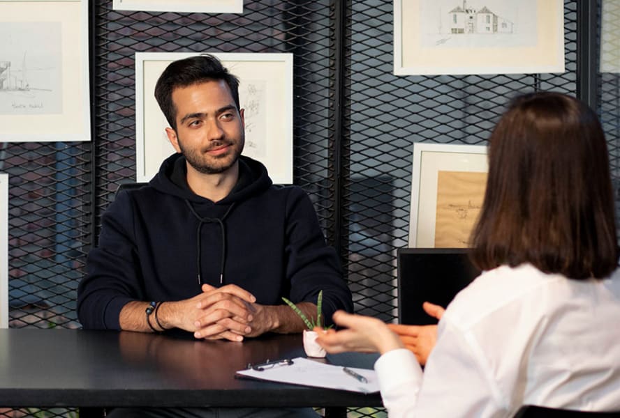 Woman in an interview with a man, he has sketches behind him on a wire mesh wall, she has paper and a laptop