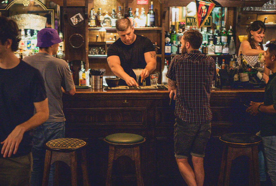 Three men stand at a pub bar while the bartender serves a cocktail