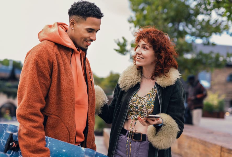 Male and female student stand outside, she holds a phone, he holds a skateboard