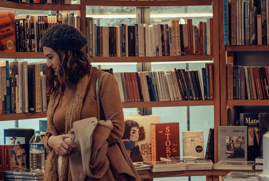 A woman in a brown jumper and scarf with a grey hat browses the shelves of a bookshop