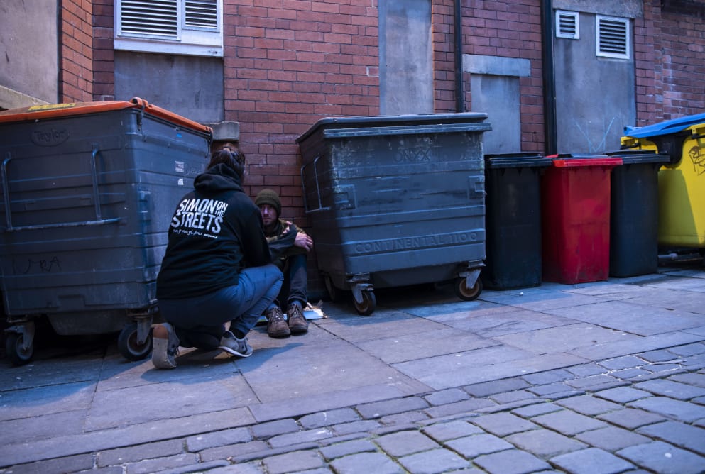 An outreach is bending down on one knee to talk to a man sat on the floor between two bins