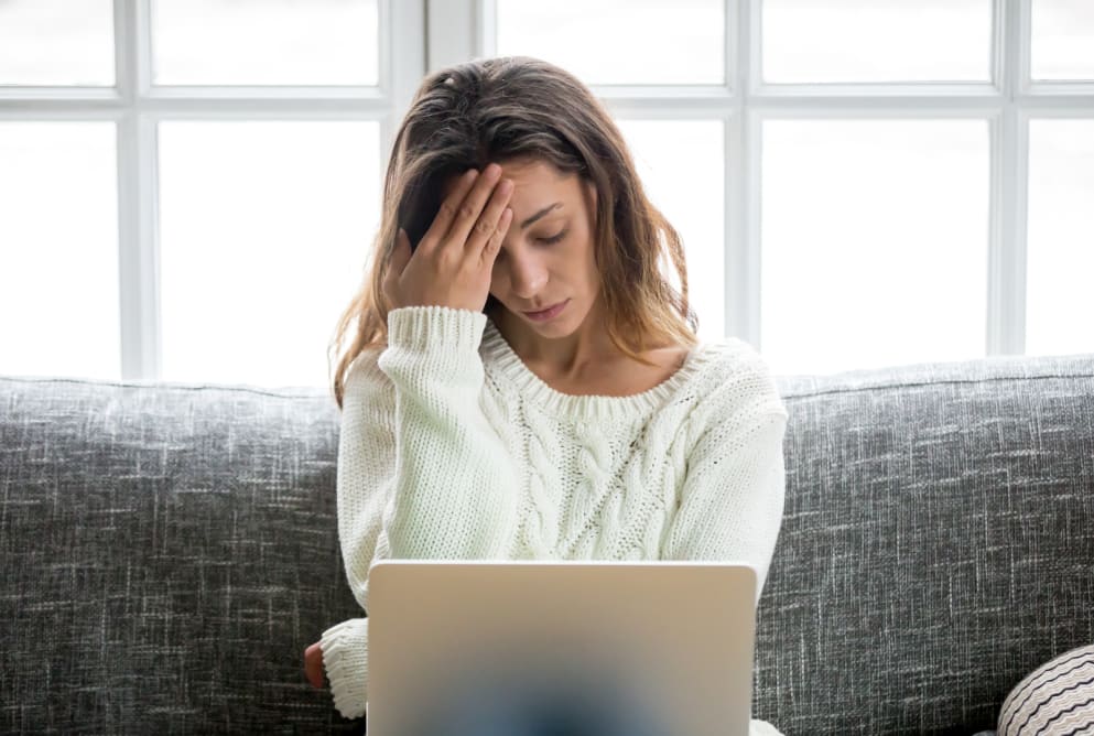 A woman puts her head to her forehead whilst staring at her laptop. She looks worried.