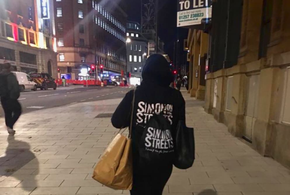 A person carries a Simon on the Streets bag on their back. They are walking down a dark street.
