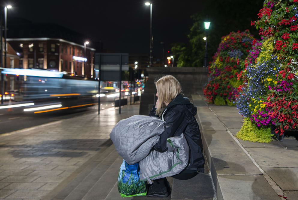 A woman sits on some steps. She has a sleeping bag in her lap. Cars speed off past her. Flowers can be seen behind her.