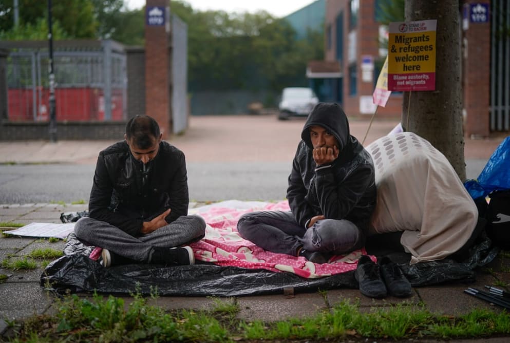 Two men sit on the floor with a blanket and plastic sheeting underneath them. One man is looking down, the other has a hood up and is resting his face on his chin.