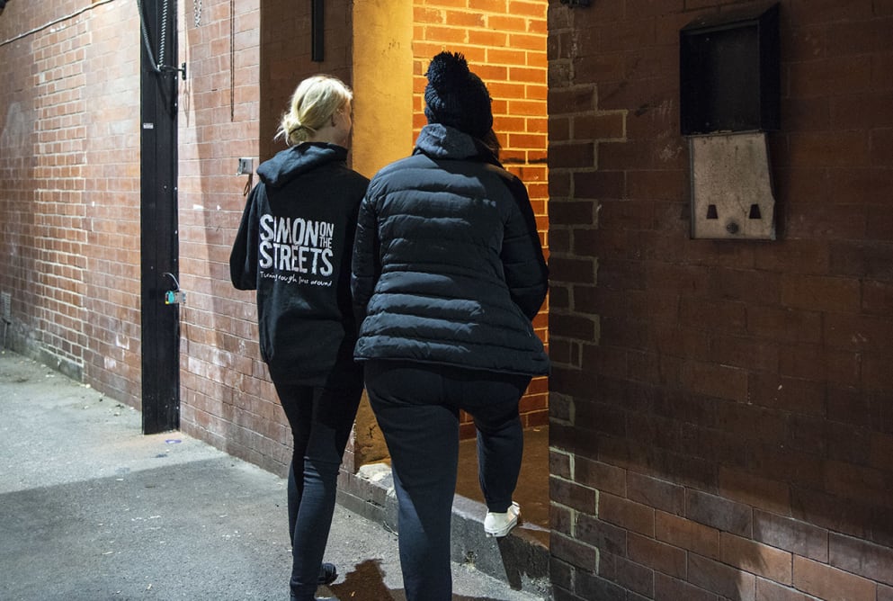 Two women walk through a doorway. The woman on the left has a Simon on the Streets hoodie on.