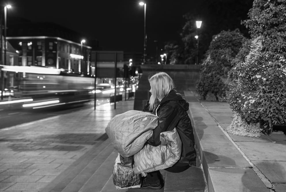 A woman sits on a set of steps. She is holding a sleeping bag and looking at the traffic passing by.