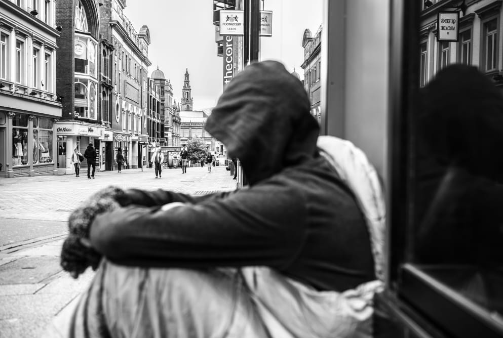 A man is sat on the ground with his hood up. He is in a sleeping bag. He gazes down the street, where buildings can be seen in the distance.