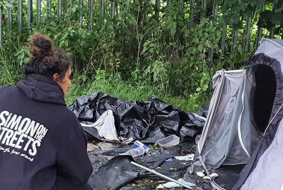 A woman is crouched next to the entrance of a tent. Her back is turned and she is wearing a Simon on the Streets hoodie.