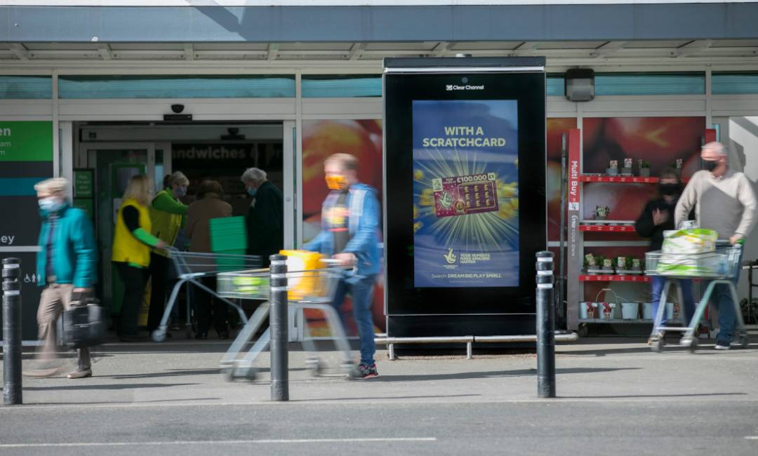 Camelot ad on digital screen outside Asda with people walking past