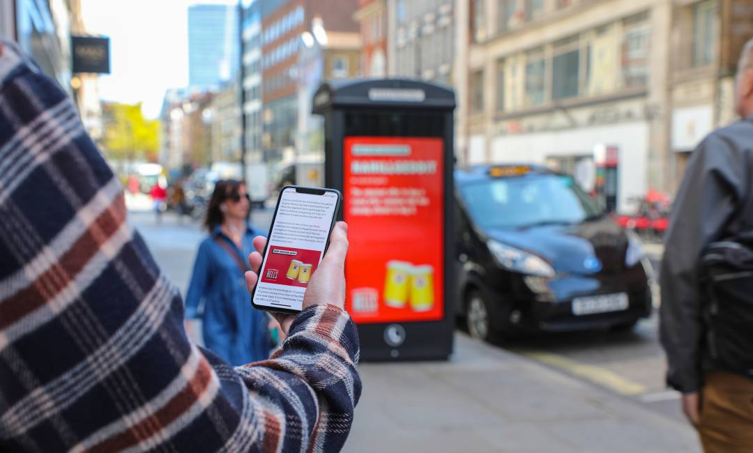 Person holding mobile phone with a beer advert on screen and a digital outdoor advertising panel with the same brand in the distance