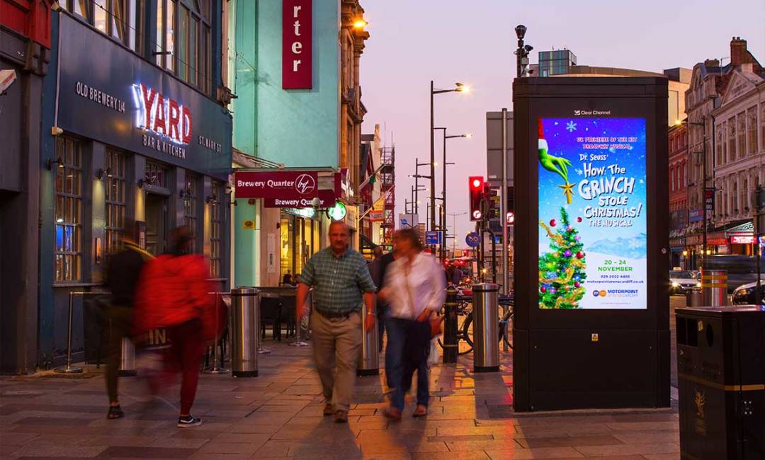 People walking past an Adshel Live display, featuring an advertisement for How the Grinch Stole Christmas, at night