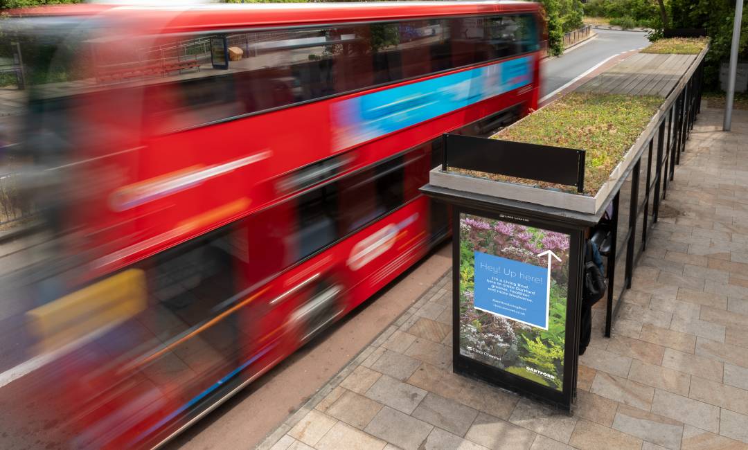 A double-decker bus drives past a Living Roof bus shelter during the day