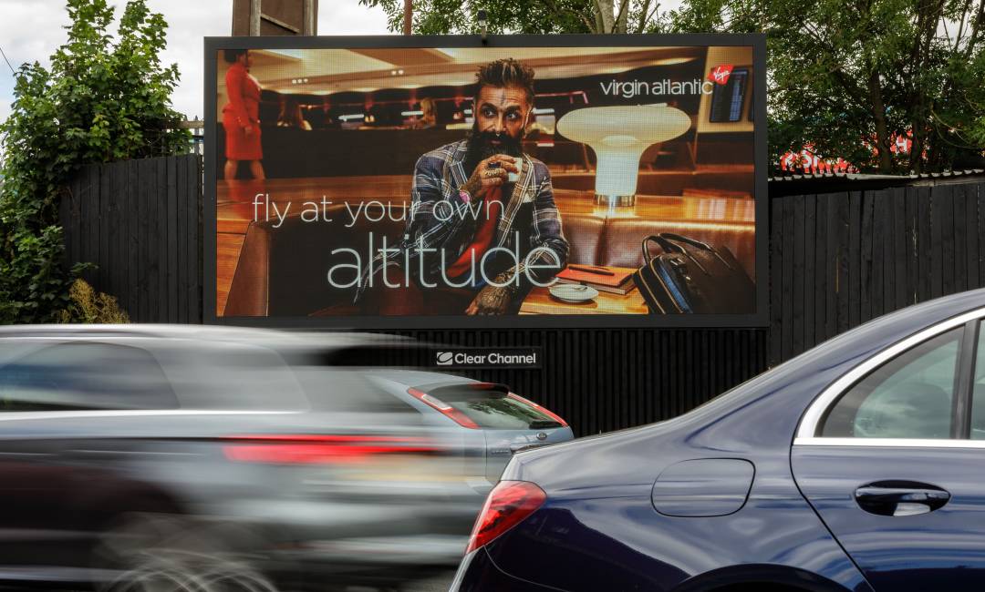 A Virgin Atlantic advertisement of a person drinking a coffee in a Virgin Atlantic airport lounge, on a Clear Channel billboard by a busy road